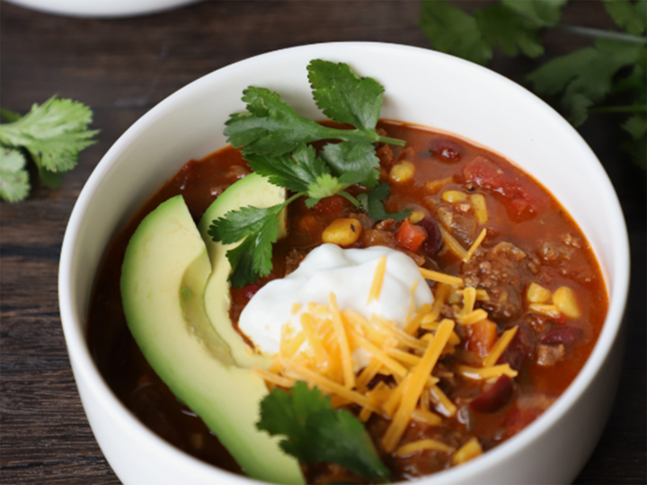 Bowl of chili with avocado slices, sour cream, shredded cheese, and cilantro garnish.